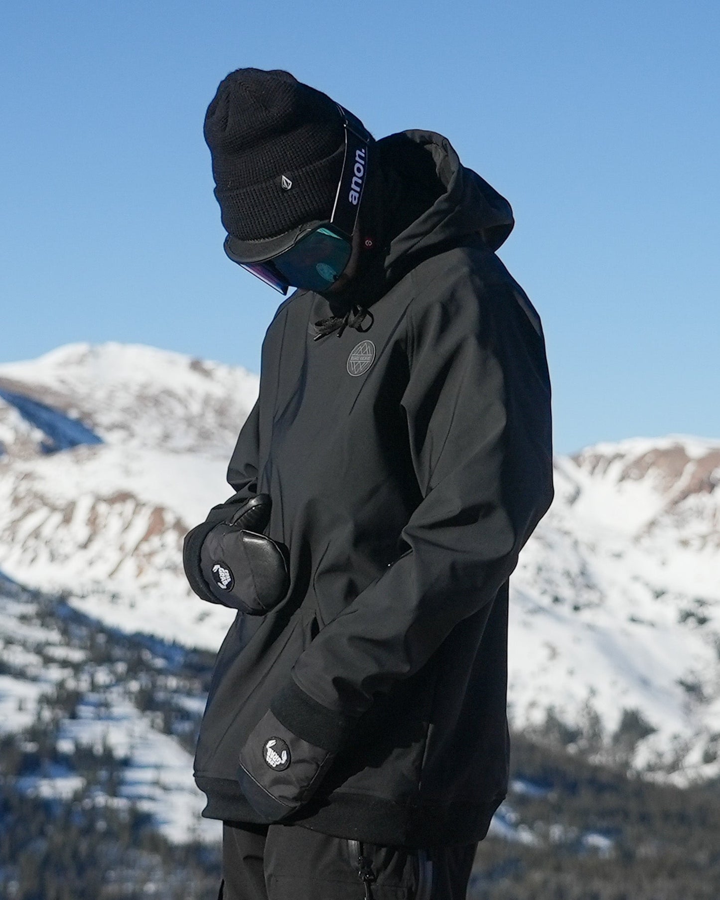 A person wearing a black waterproof riding hoodie with a hood, standing against a snowy mountain background. The hoodie has a kangaroo pocket, mesh pit vents, and zippers.